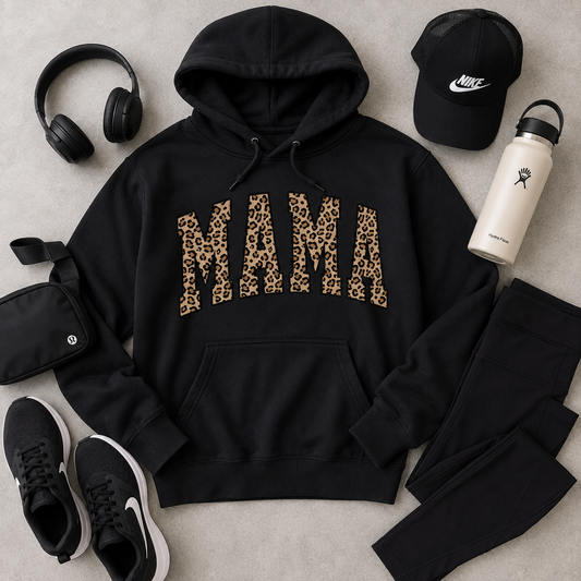 A black pullover hoodie with the word 'MAMA' printed across the chest in leopard-print letters, styled in a flat lay with a black cap, white water bottle, black headphones, black crossbody bag, black sneakers, and black leggings on a light gray surface.
