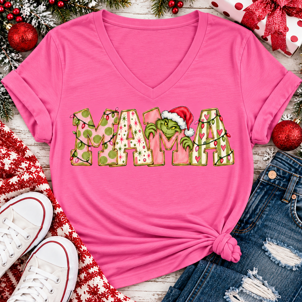 Christmas MAMA graphic shirt featuring pink and green patterned lettering, colorful holiday lights, and a Grinch-inspired character wearing a Santa hat.